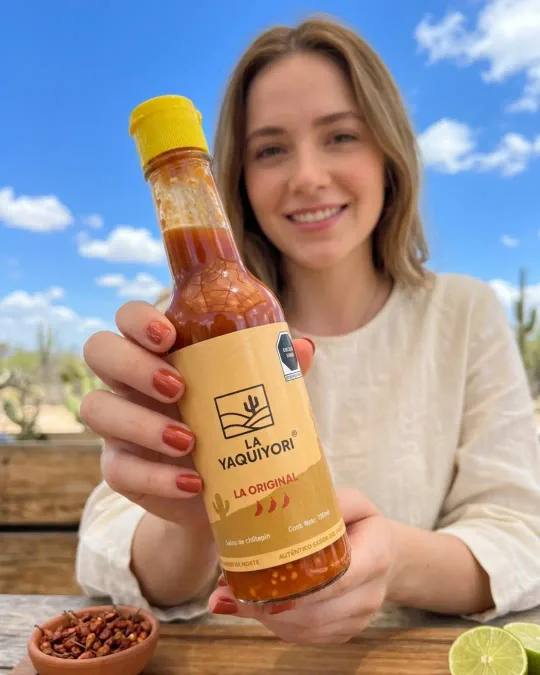Una mujer sostiene una botella de salsa La Original de chiltepín silvestre La Yaquiyori, junto a un cuenco de barro con chiltepines secos y limón fresco, con el desierto de Sonora al fondo